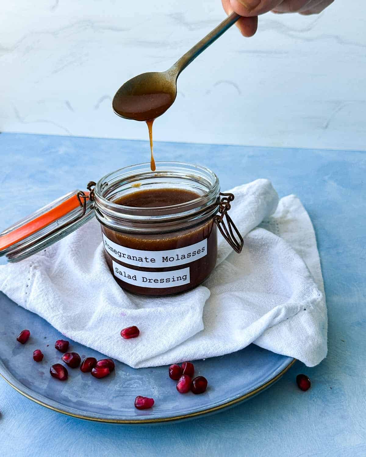 Spoon drizzling thick pomegranate molasses dressing back into a labeled glass jar on a blue plate.