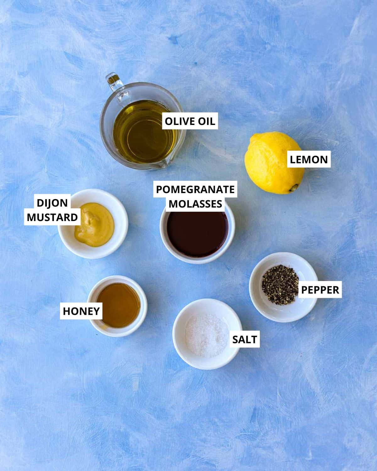 Overhead ingredient image for pomegranate molasses dressing with olive oil, lemon, Dijon mustard, honey, salt, and pepper.