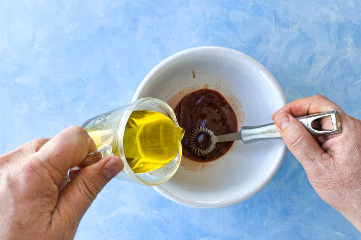 Olive oil being poured into a bowl of pomegranate molasses dressing while whisking.