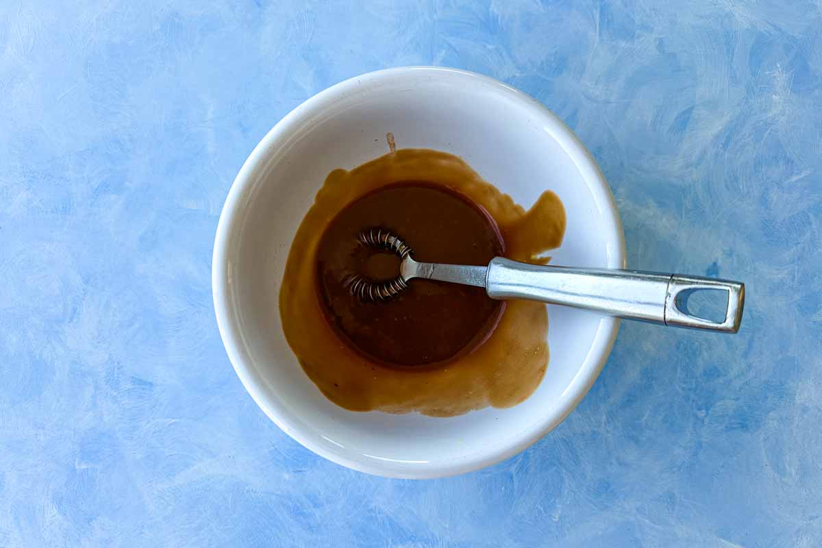 Overhead view of whisked pomegranate molasses dressing in a white bowl with a small whisk.