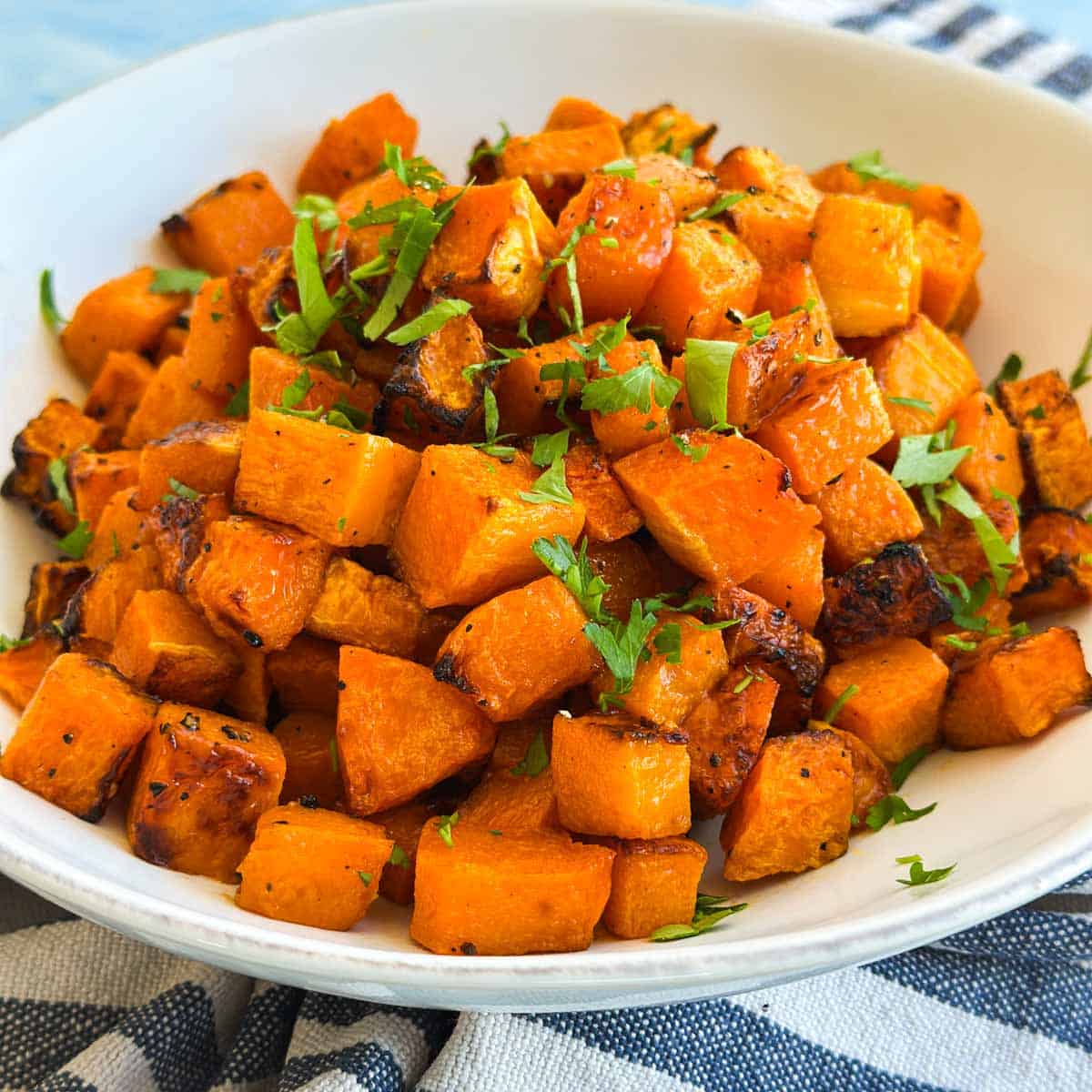 Close-up of roasted butternut squash cubes with caramelized edges and chopped parsley in a white bowl.