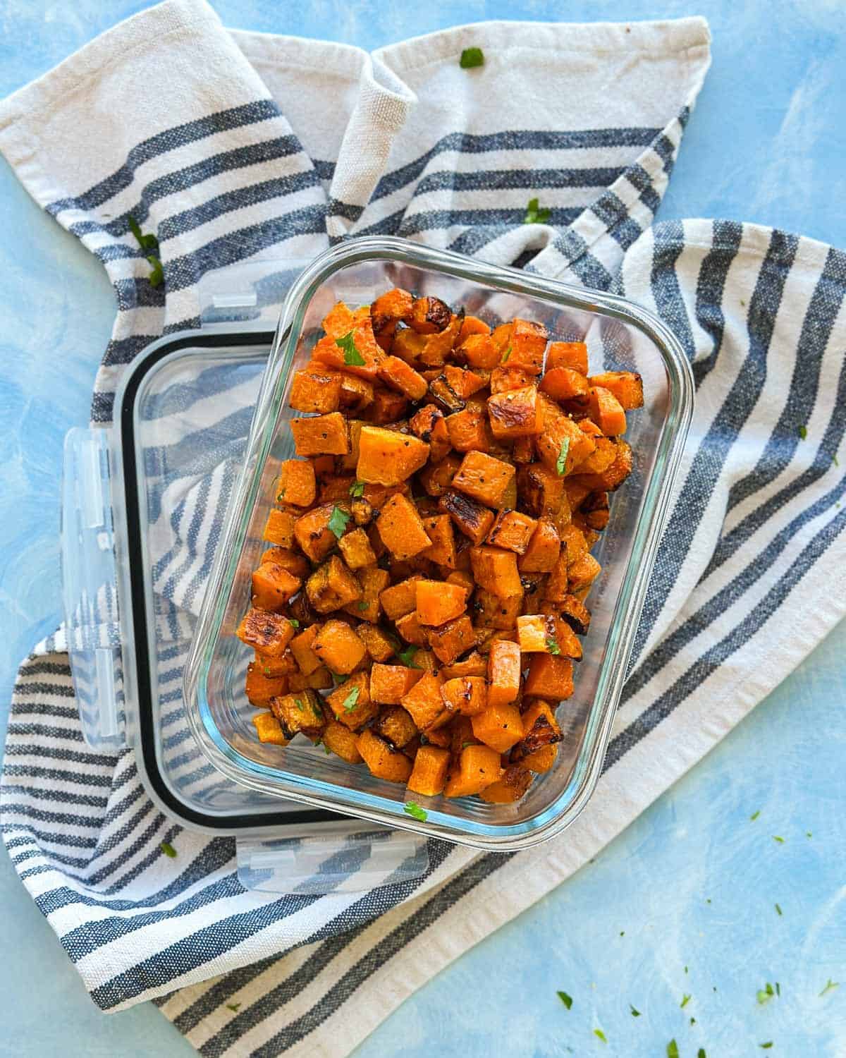 Roasted butternut squash cubes stored in a rectangular glass container, styled on a striped kitchen towel for meal prep.