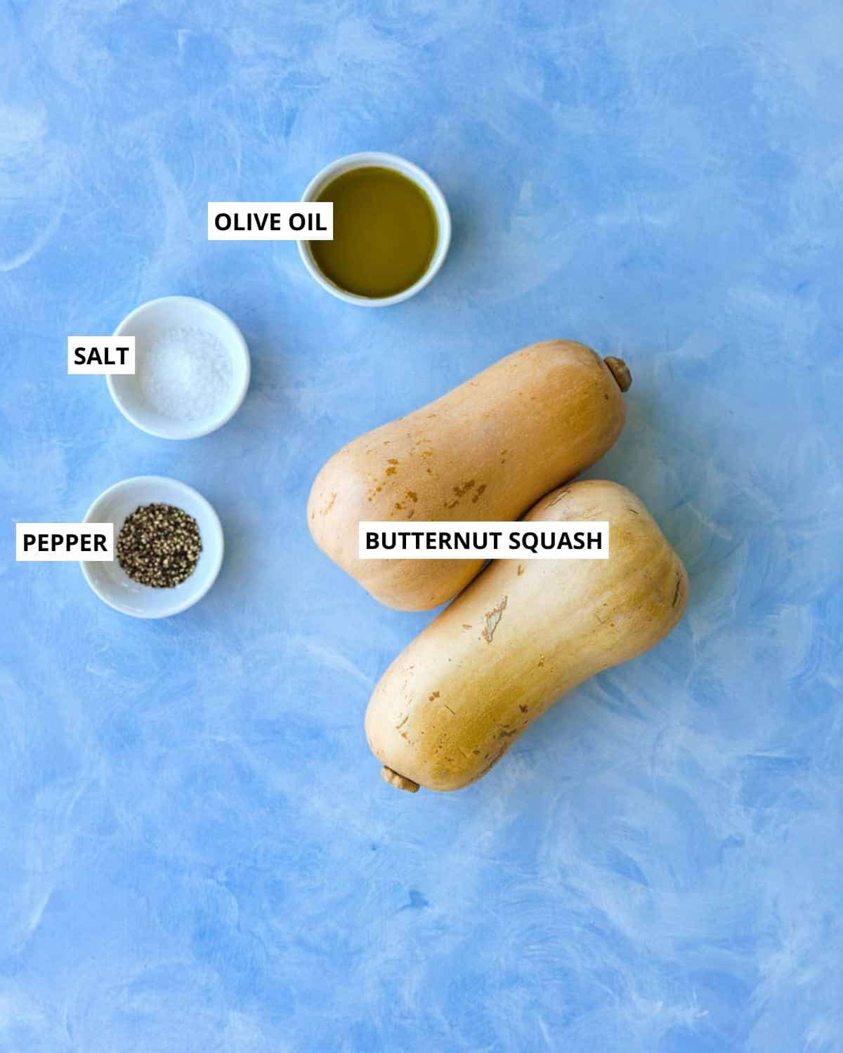 Ingredients for roasted butternut squash, including butternut squash, olive oil, salt, and pepper on a blue background.