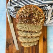 Overhead of a seed-crusted no-knead loaf, half sliced, on a wooden board with a bread knife and striped towel.