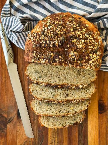 Overhead of a seed-crusted no-knead loaf, half sliced, on a wooden board with a bread knife and striped towel.