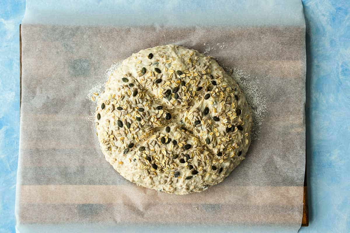 Scored dough topped with oats and seeds, ready to bake in a parchment-lined Dutch oven.