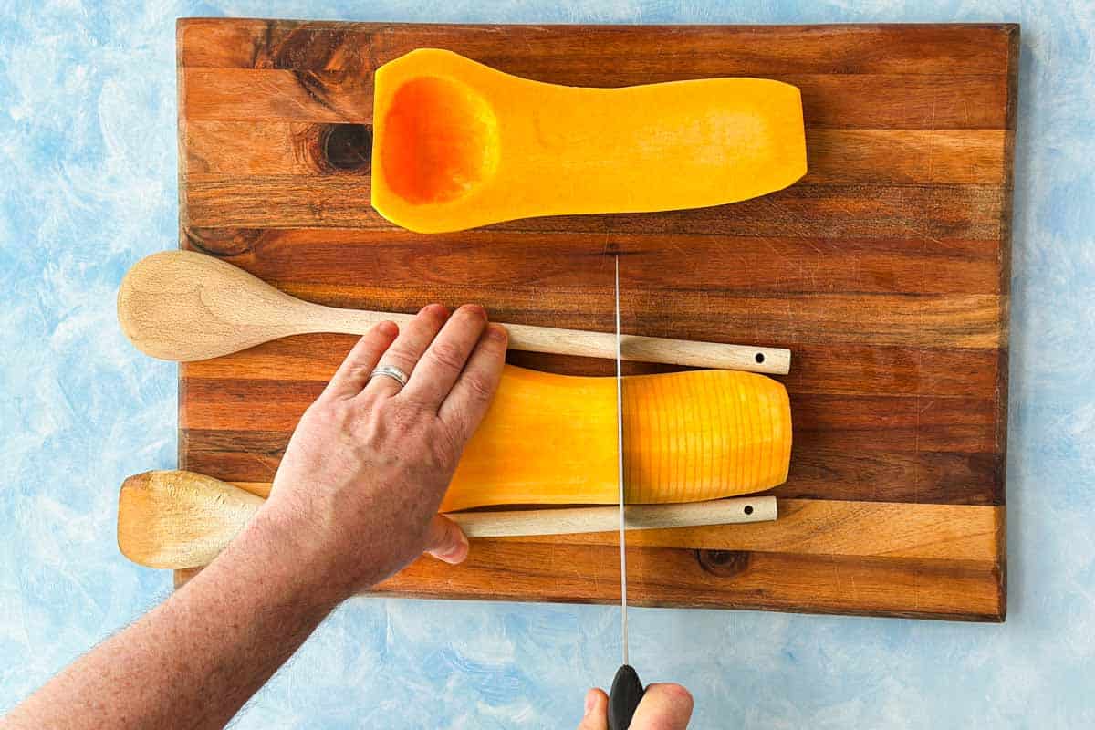 Butternut squash being carefully sliced hasselback-style between two wooden spoon handles on a cutting board.