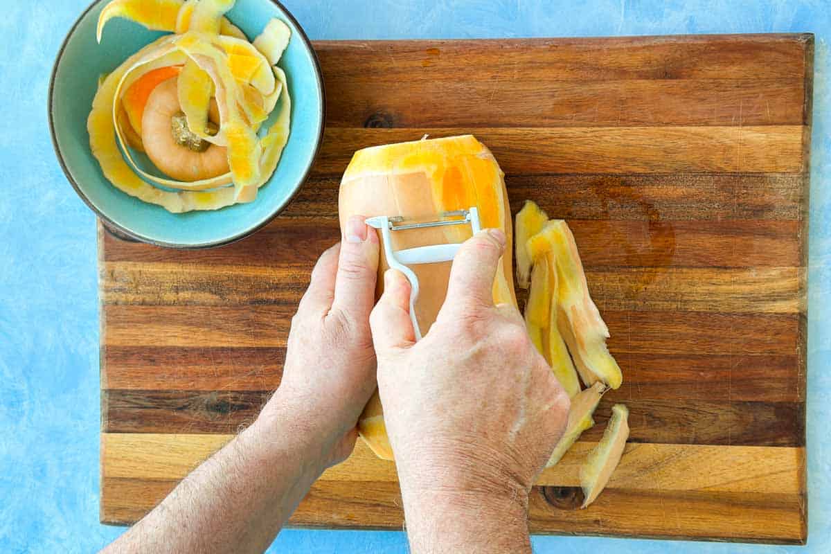Top-down step-by-step photo of a butternut squash being peeled with a vegetable peeler, with the peelings collected in a bowl.