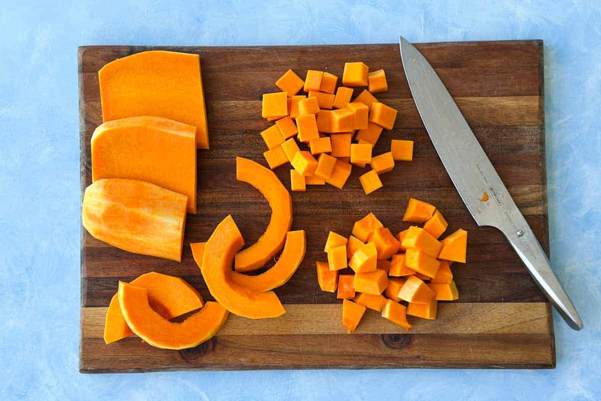 Prepared butternut squash cut into chunks, slices, and cubes on a wooden cutting board with a chef’s knife.