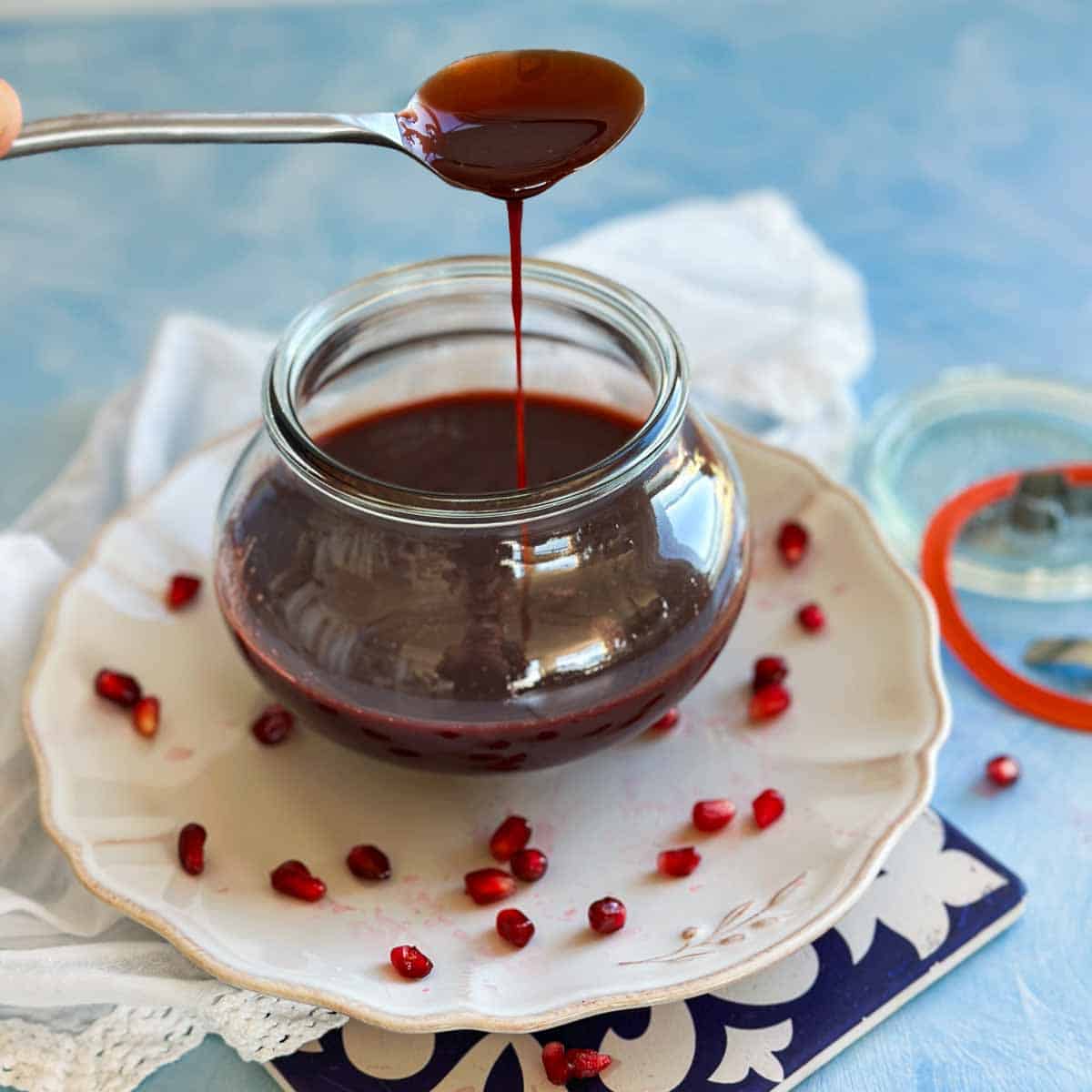 Thick homemade pomegranate molasses dripping from a spoon back into a glass jar, with pomegranate seeds scattered on the plate.