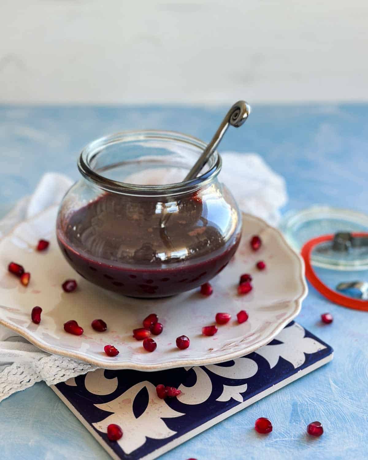 Glass jar of homemade pomegranate molasses with a spoon, styled on a white plate with pomegranate seeds.