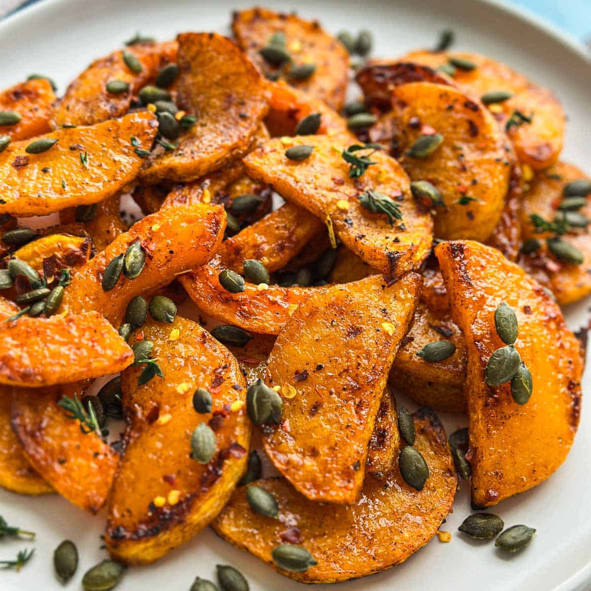 Tight close-up of golden roasted butternut squash slices with a glossy finish, scattered pumpkin seeds, and thyme.