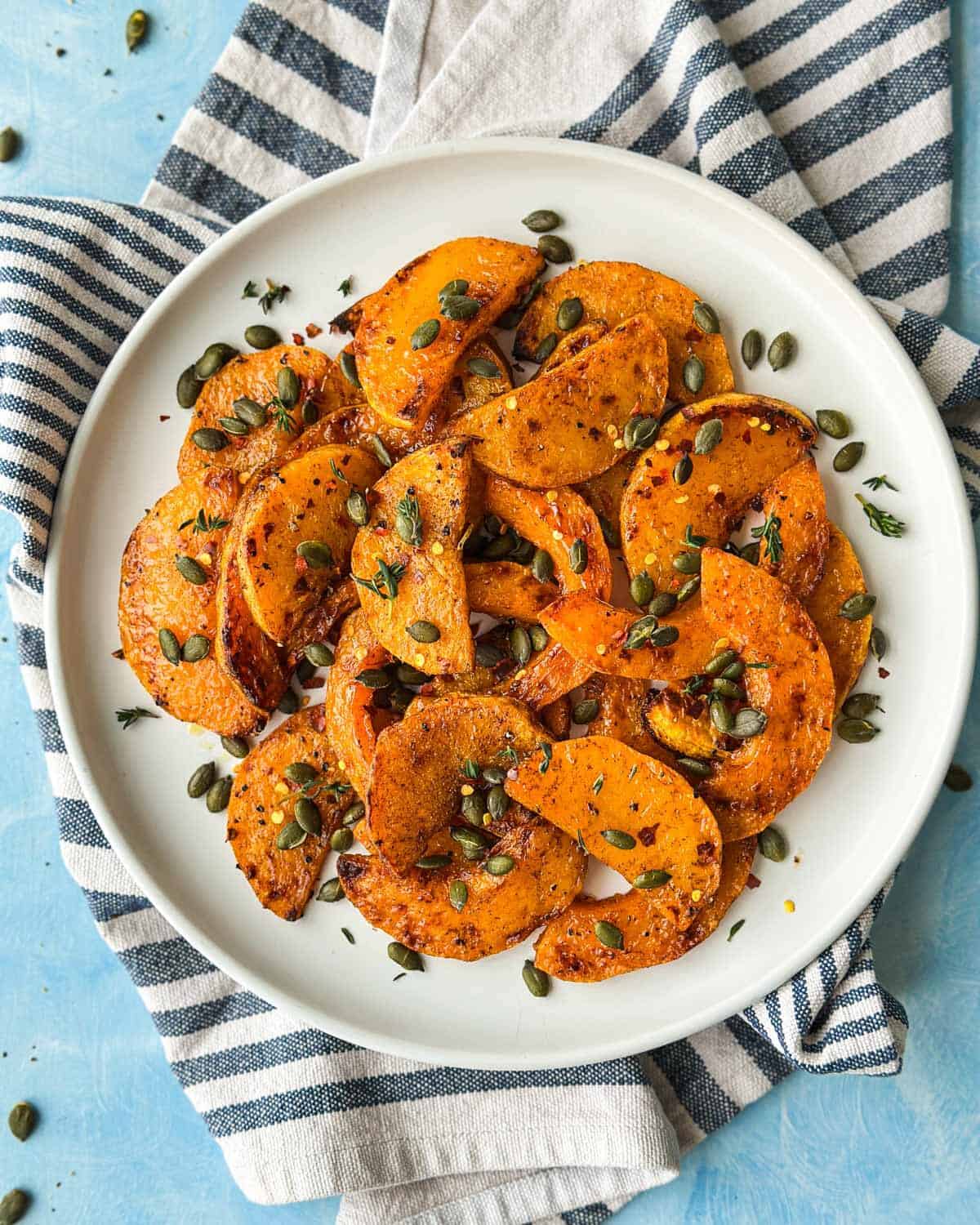 Overhead shot of cinnamon roasted butternut squash slices arranged on a white plate with pumpkin seeds and thyme.