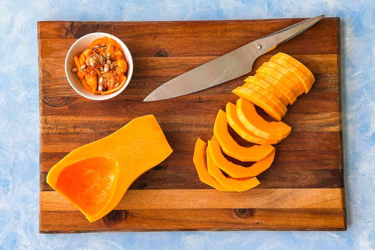 Butternut squash sliced into half moons on a wooden cutting board with the seeds removed and a chef’s knife nearby.