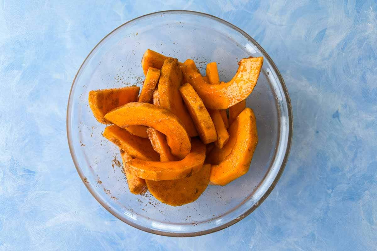 Butternut squash slices in a glass bowl coated with oil and seasonings before roasting.