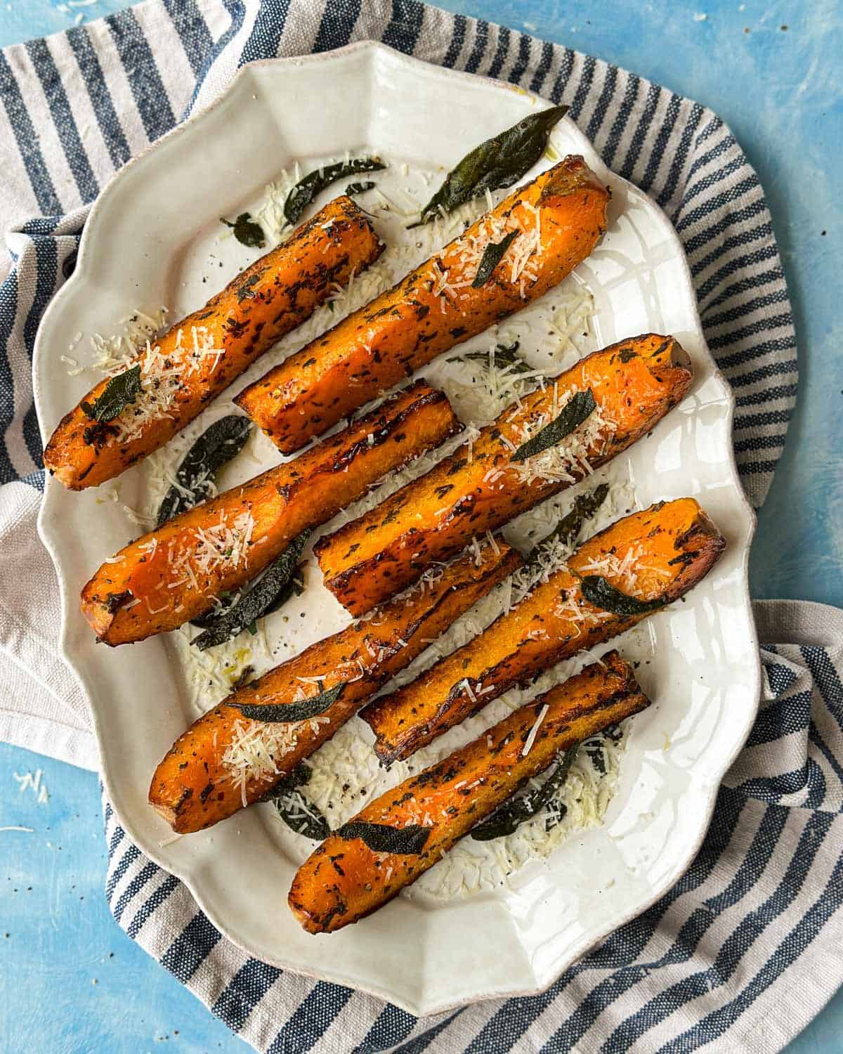 Overhead view of roasted butternut squash wedges sprinkled with Parmesan cheese and crispy sage leaves on a white platter.