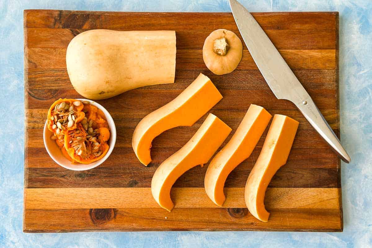 Butternut squash cut into long wedges on a wooden cutting board with the seeds removed and a chef’s knife nearby.