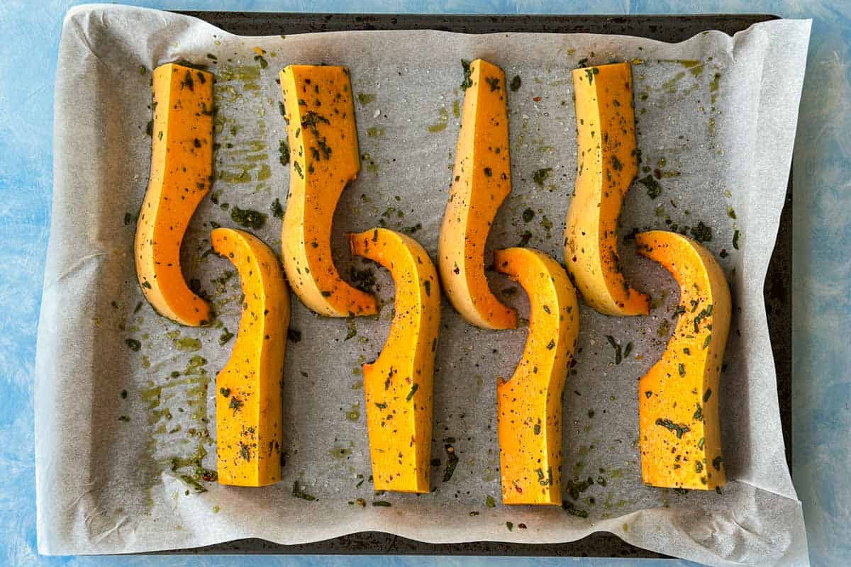 Raw butternut squash wedges seasoned with herbs, olive oil, salt, and pepper on a parchment-lined baking sheet before roasting.