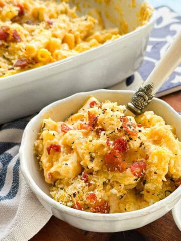 Close-up of a scalloped white bowl of butternut mac and cheese with bacon and breadcrumbs, a spoon resting in the pasta.