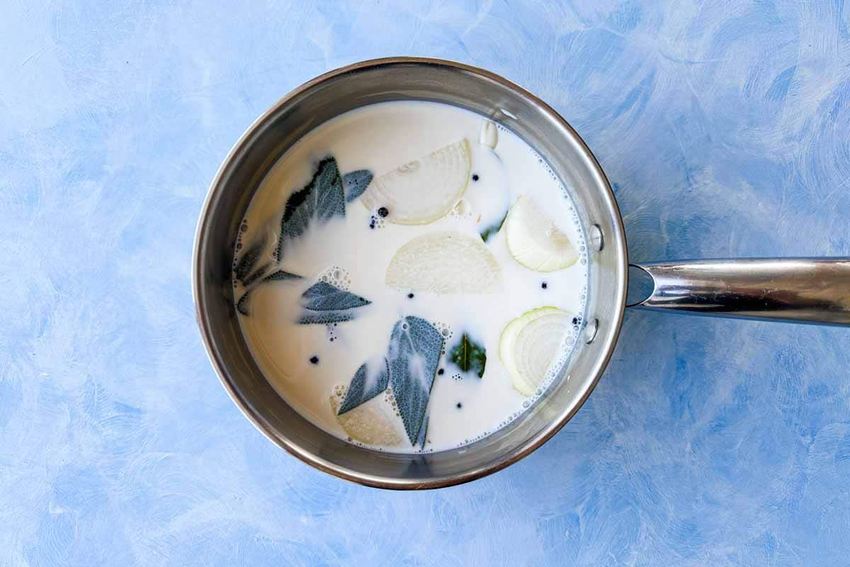 Milk infusing with onion wedges, sage leaves, bay leaf, and peppercorns in a stainless steel saucepan.