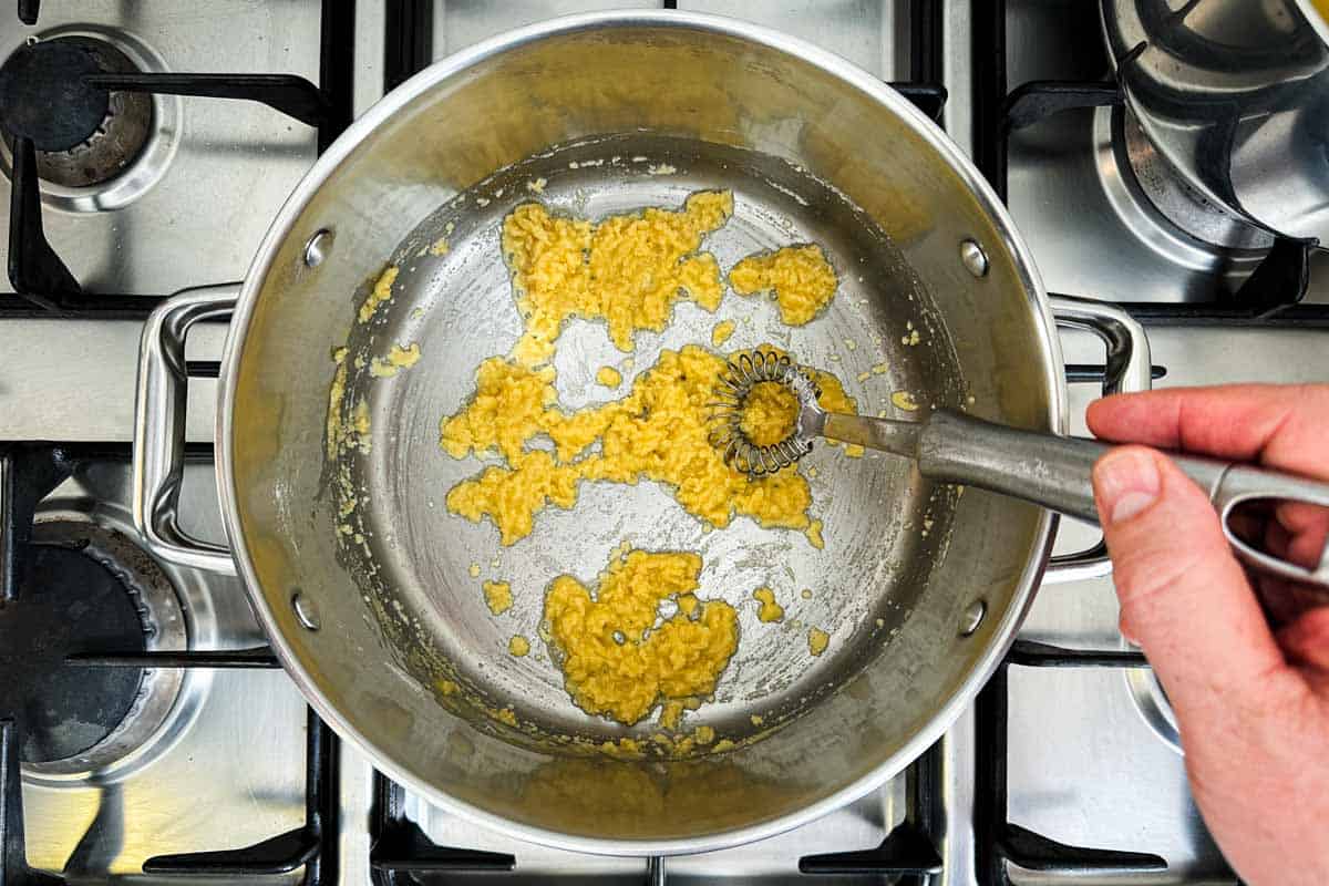 A hand whisking a yellow butter and flour roux in a stainless steel pot on the stove.