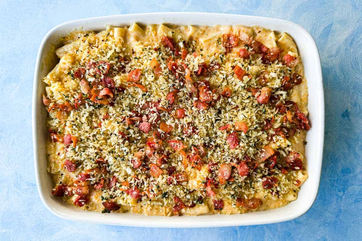 Unbaked butternut mac and cheese in a white dish, covered with herbed panko breadcrumbs and raw bacon pieces.