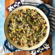 Overhead shot of brown and wild rice pilaf studded with apples, cranberries, and pecans in a blue Dutch oven on a wood board.