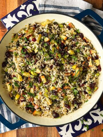 Overhead shot of brown and wild rice pilaf studded with apples, cranberries, and pecans in a blue Dutch oven on a wood board.