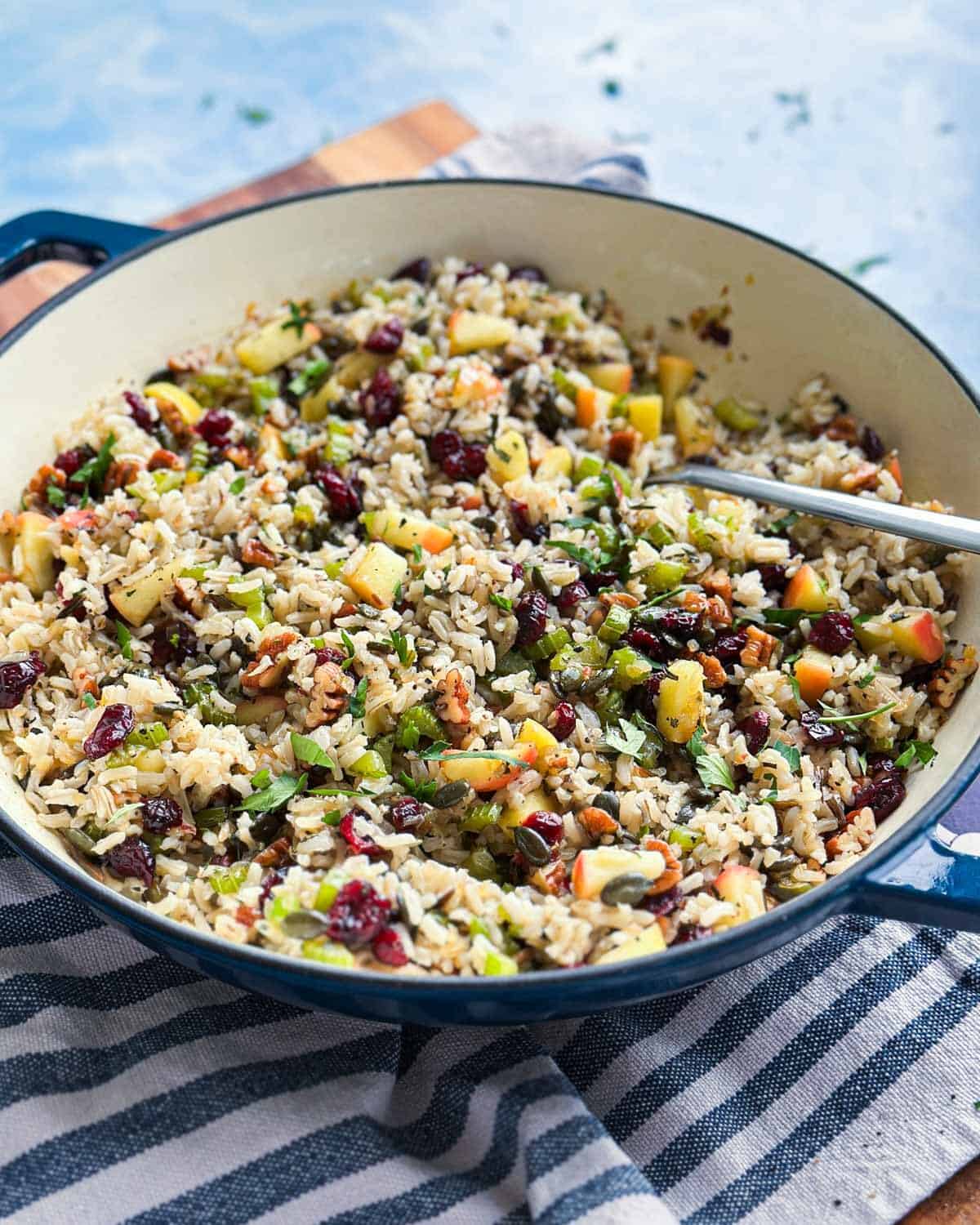 Vertical shot of the pilaf in its pot with a spoon inside, resting on a wood board beside a patterned tile and striped towel.