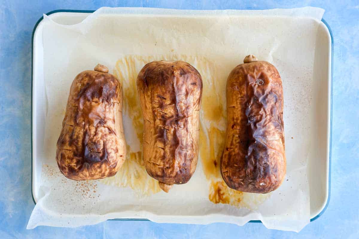 Three whole butternut squash after roasting, skins wrinkled and charred, with caramelized juices on the parchment.