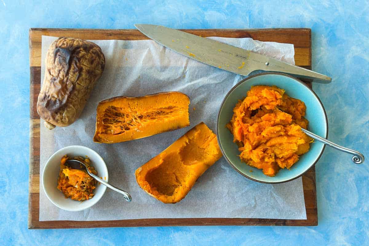 Roasted butternut squash halved on a cutting board, with seeds scooped into a small bowl and the flesh mashed in a blue bowl.